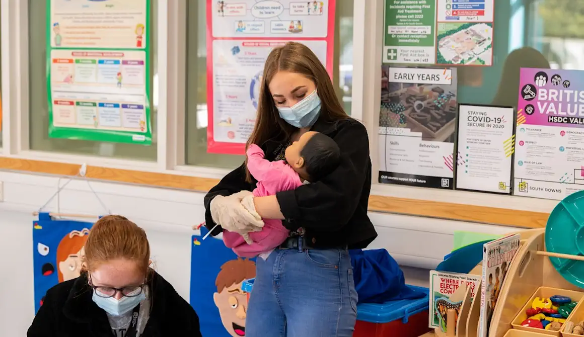 Two students, one holding a baby simulator in her arms while wearing gloves and a mask.