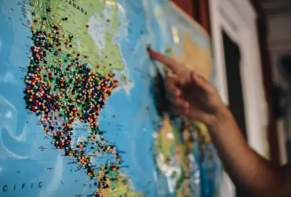 A hand points at a map of the world with pins in it.