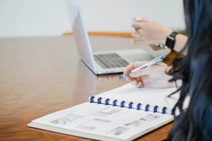 a person writing in a notebook at a desk with someone else with a laptop