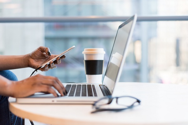 a woman typing at a laptop whilst using her mobile phone with a coffee