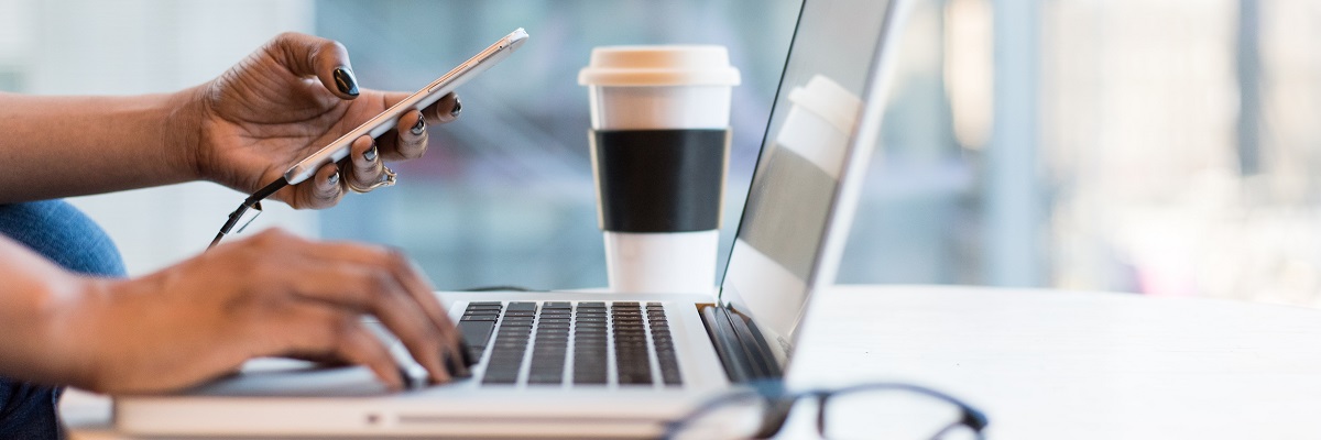 a woman typing at a laptop whilst using her mobile phone with a coffee