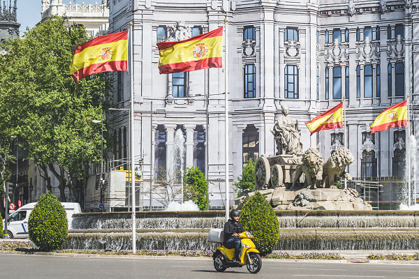 a man riding a moped with spanish flags flying behind him in front of a building