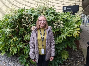 Caroline is smiling and stood in front of a bush