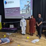 Three people standing next to a crime scene dummy, one of the people is dressed in a full white CSI outfit