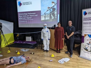 Three people standing next to a crime scene dummy, one of the people is dressed in a full white CSI outfit
