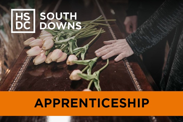 A closeup of hands and flowers on a wooden coffin.