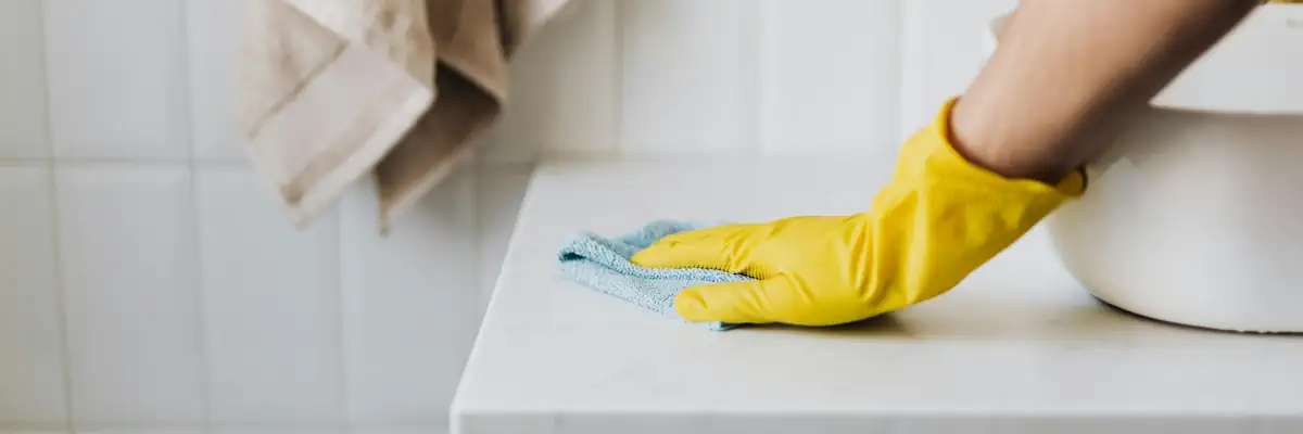 Closeup of a yellow gloved hand cleaning a white bathroom counter with a cloth.