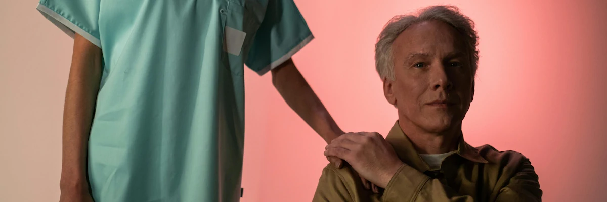 An older gentleman sits, grasping a nurse's hand on his shoulder.