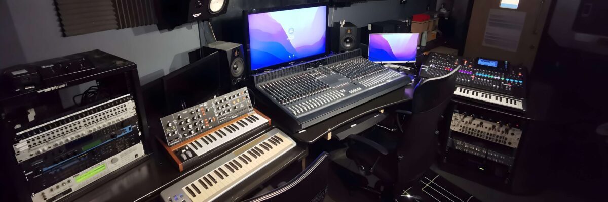 A photo of studio A, including two monitors, a large mixing desk, several stereo monitor speakers, midi keyboards and synthesizers as well as an analogue plugin rack.
