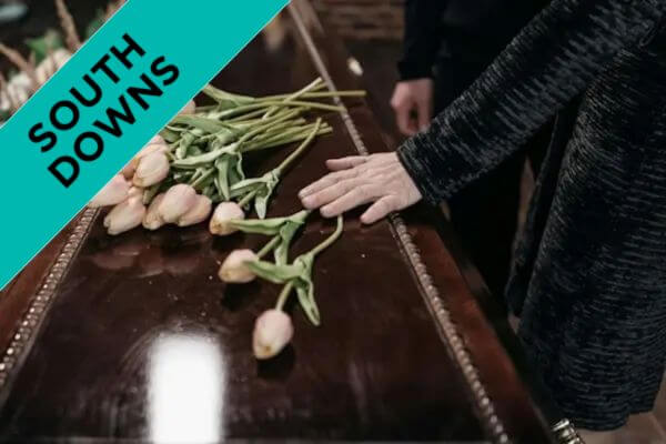 A closeup of hands and flowers on a wooden coffin.